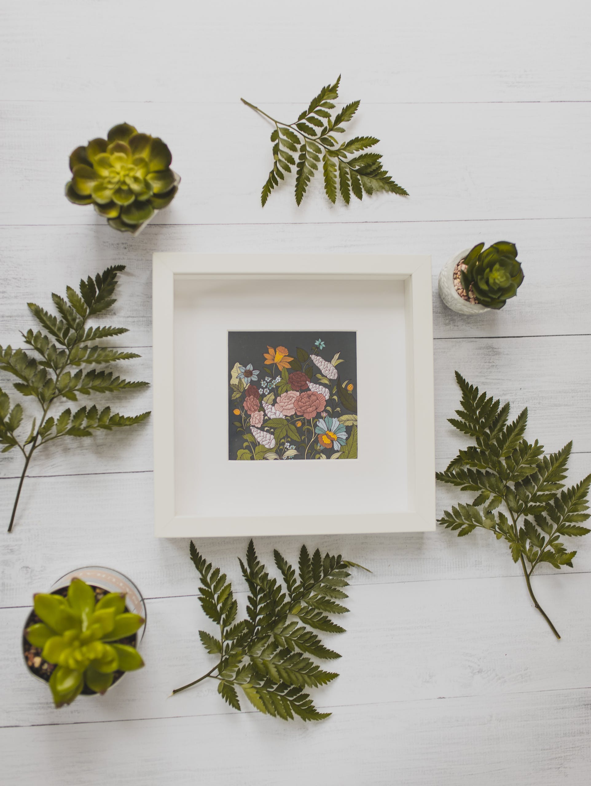 Top view composition of white square frame with floral picture placed on white wooden desk amidst green lush succulents and plant leaves